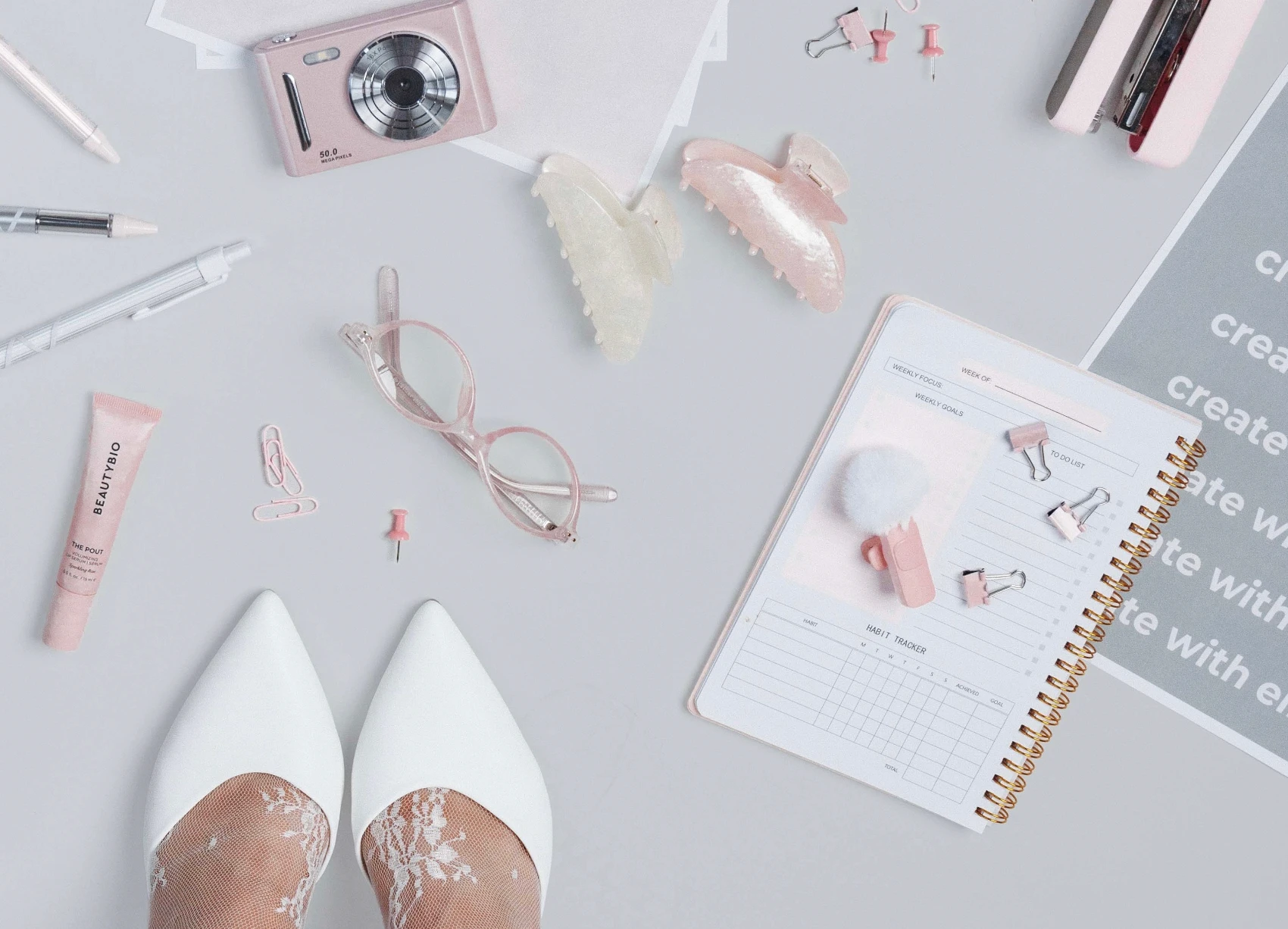 Pink and white desk flat lay with a camera, glasses, hair clips and a planner